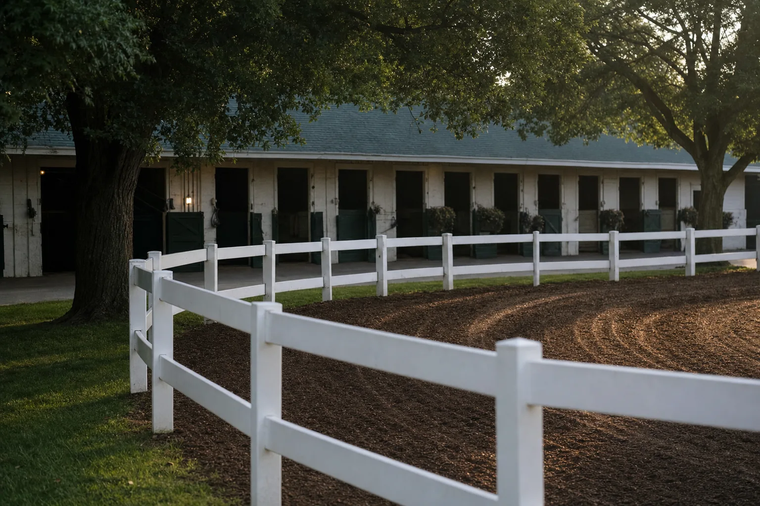Empty paddock walking ring with white post-and-rail fence and saddling-stall barn in soft morning light.